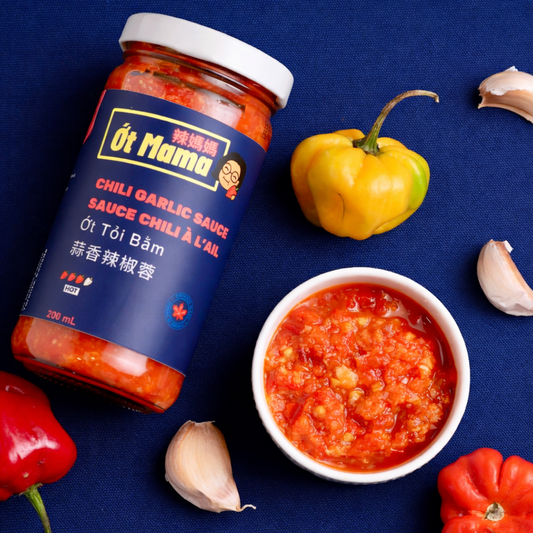 A jar of Ót Mama Chili Garlic Sauce is arranged on a dark blue fabric background with whole and peeled garlic cloves, red and yellow bell peppers, and a small white bowl filled with the thick red sauce.